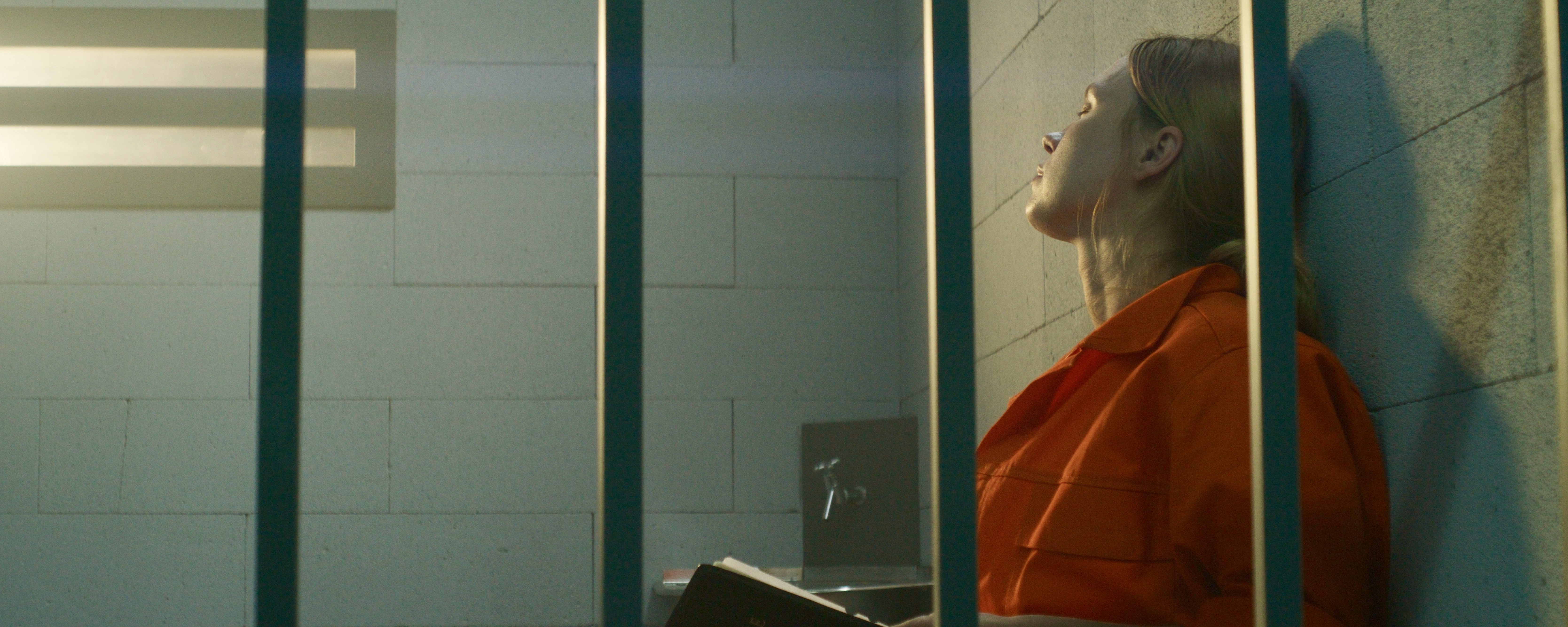 Female prisoner in orange uniform sits on bed behind metal bars